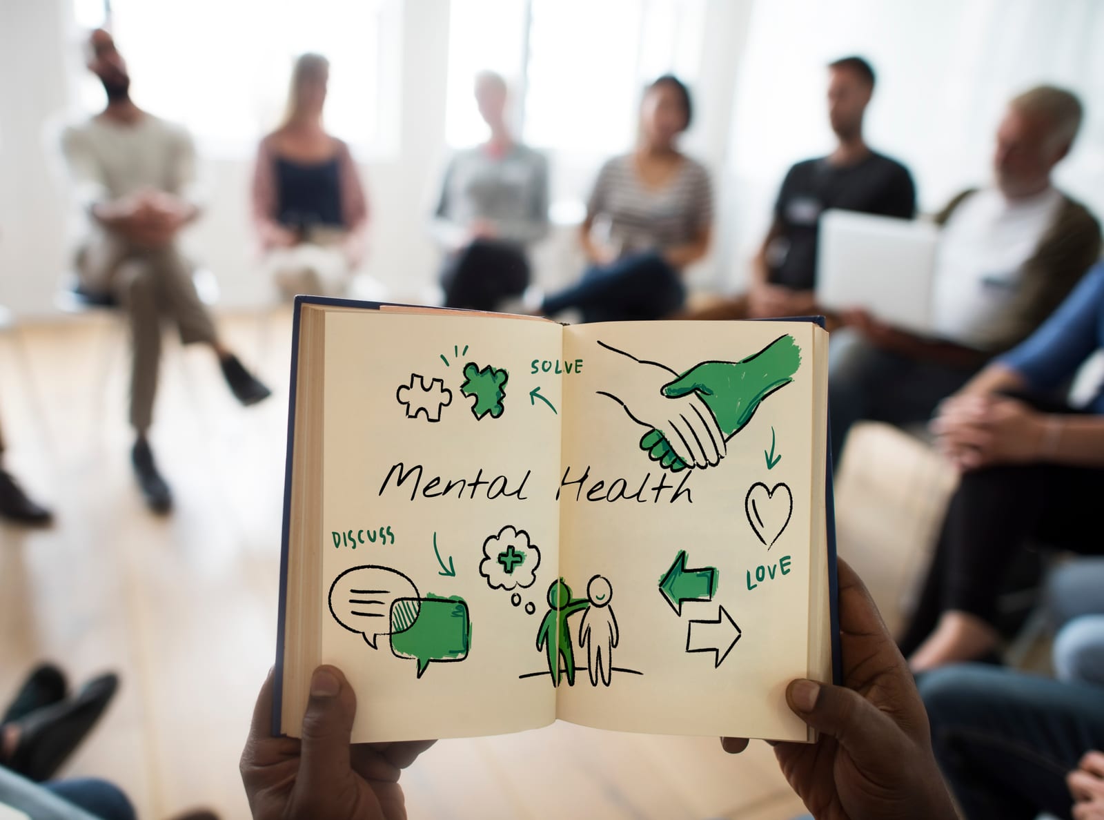 An open notebook of mental health sketches held in front of a group therapy circle